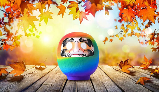 A rainbow daruma doll with both eyes left unpainted, standing on weathered wood among autumn maple leaves